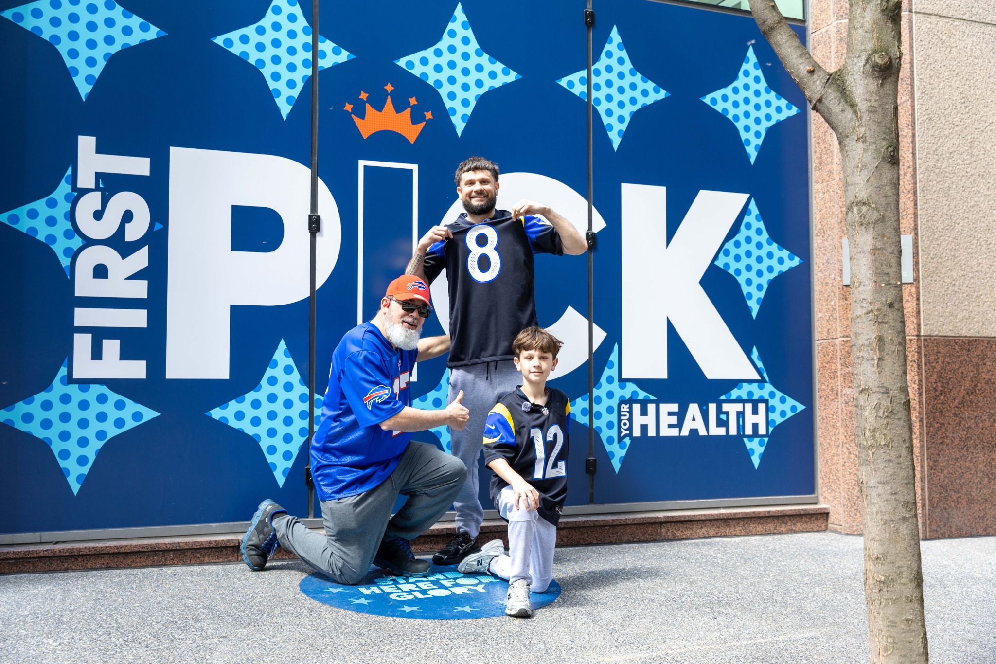 Three football fans posing in front of the First Pick Your Health sign